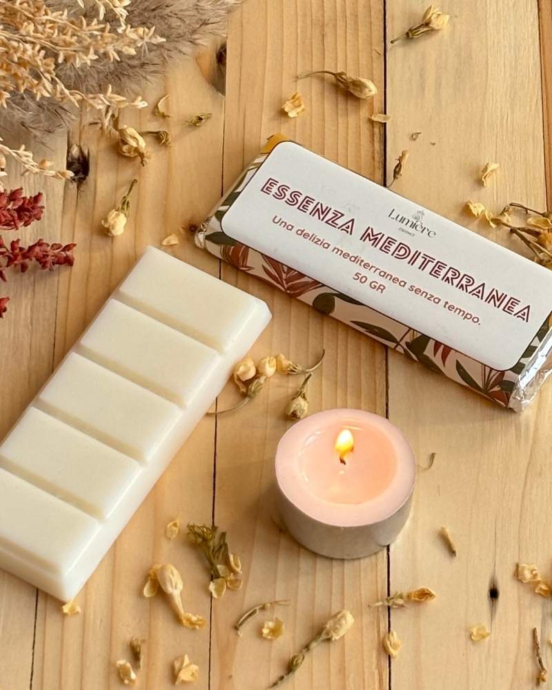 barretta in cera di cocco Lumiere Essence  Essenza Mediterranea con una piccola candela affianco accesa su piano di legno e piccoli fioi attorno