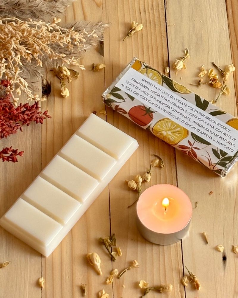 Barretta Essenza mediterranea Lumiere Essence con etichetta retro visibile una candelina accesa affianco e fiori decorativi