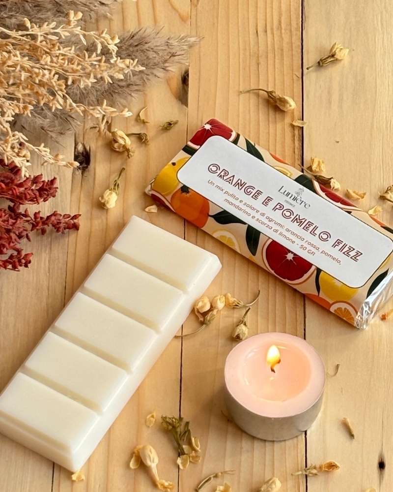 barretta di cera di cocco orange e pomelo fizz di lumière essence on piccola cadela accesa affianco e piccoli fiori decorativi su piano di legno
