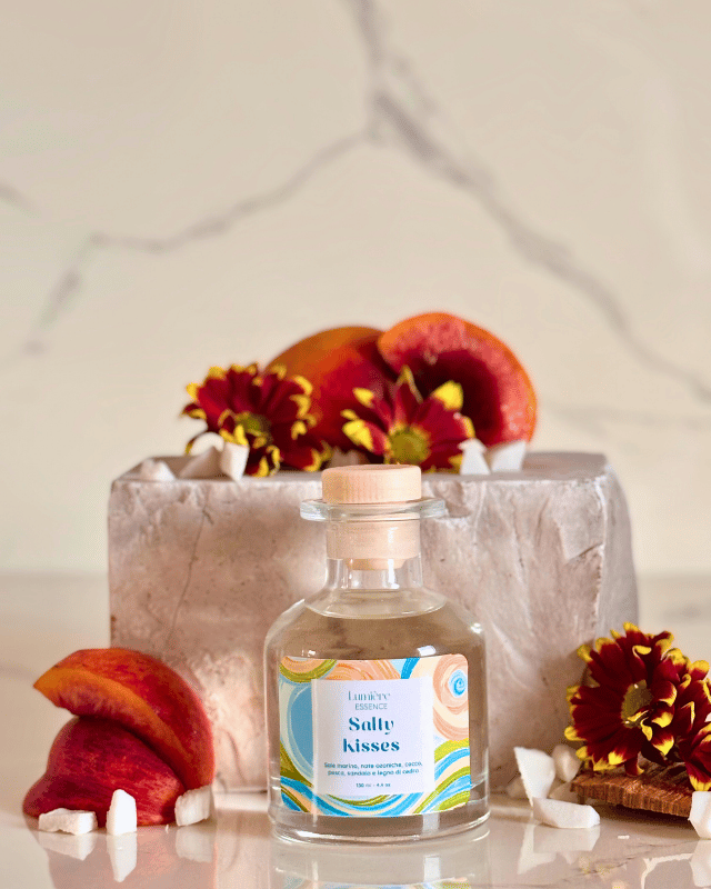 Diffusore a bastoncino in bottiglietta di vetro sullo sfondo pesche e scaglie di cocco e una pietra di marmo