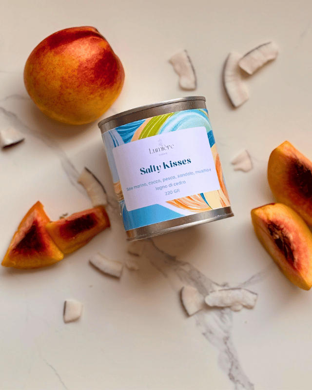 Candela Salty Kisses ripesa dall'alto realizzata in cera di cocco in barattolo di latta sullo sfondo pesche e scaglie di cocco che richiamano la fragranza